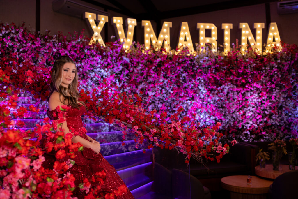 Debutante em vestido vermelho diante do muro de flores e do letreiro luminoso XV Marina