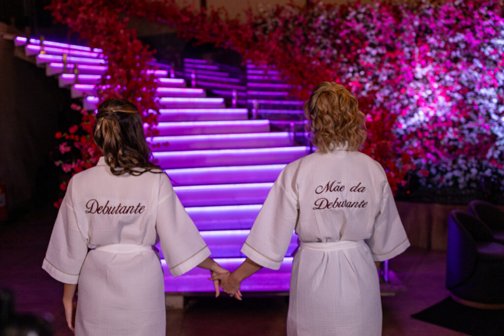 Debutante e a mãe de costas, com robes personalizados, diante da escadaria iluminada