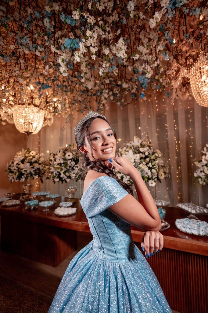 Debutante em vestido azul apoiada na mesa do bolo sob arranjos suspensos