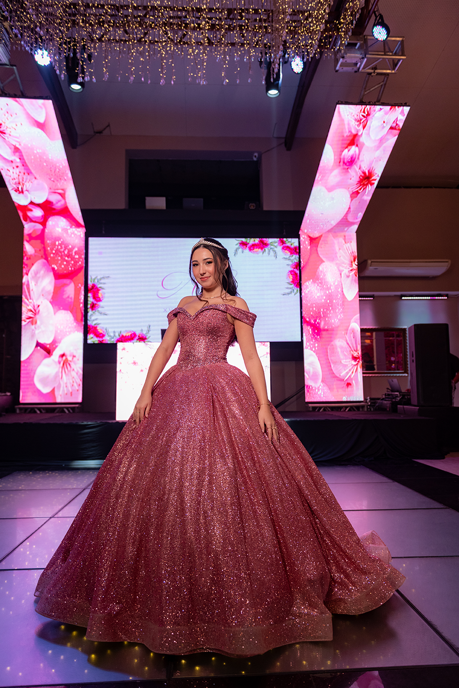 Debutante em vestido rosé diante do painel de LED com o monograma da festa