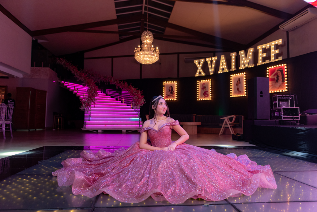 Debutante sentada com o vestido rosé espalhado diante do letreiro luminoso XV Aimée