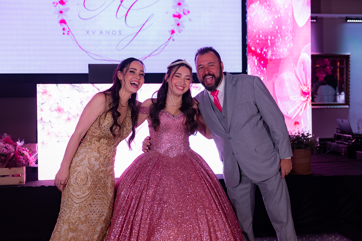 Aimée entre os pais diante do painel com o monograma da celebração