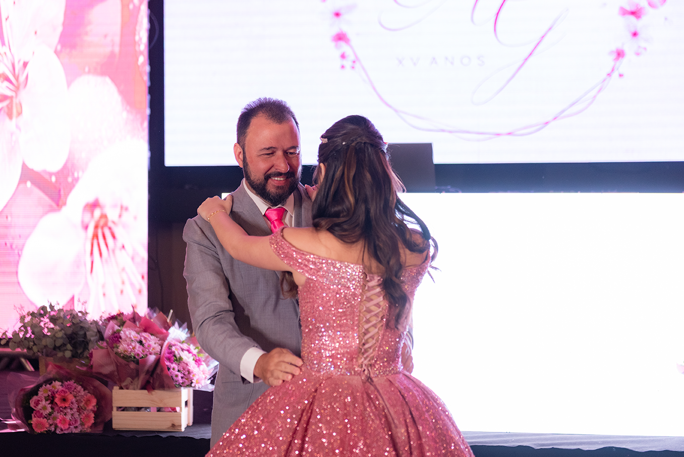 Debutante abraçada ao pai diante do painel com o monograma da festa