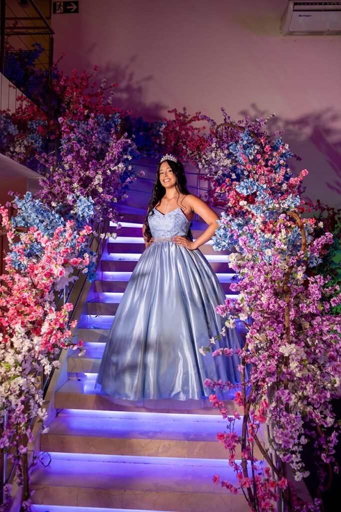 Debutante em vestido azul na escadaria decorada com flores em tons de rosa