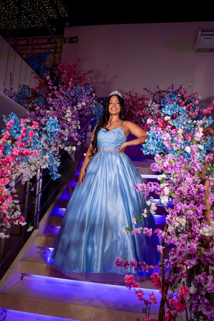 Debutante em vestido azul ao pé da escadaria decorada com flores