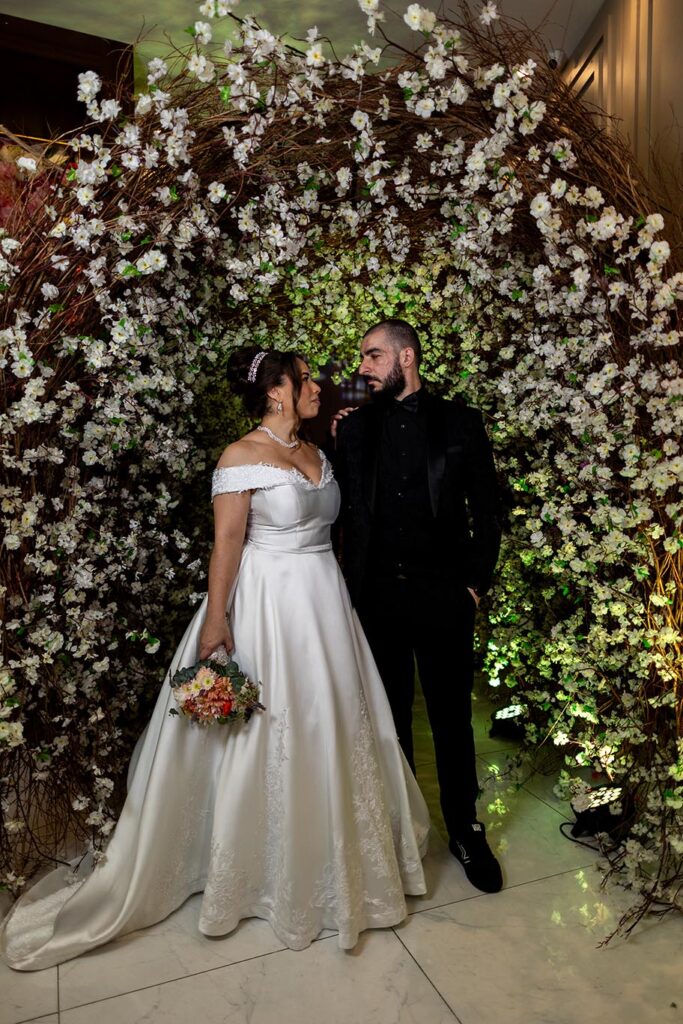Noivos posam diante da parede de flores brancas e folhagens