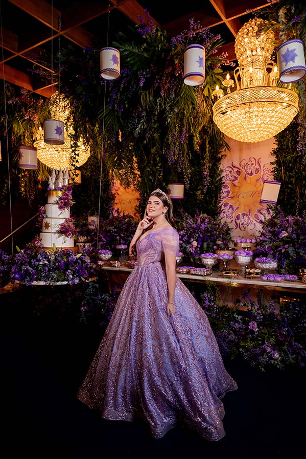 Debutante em vestido lilás ao lado da mesa do bolo decorada com flores