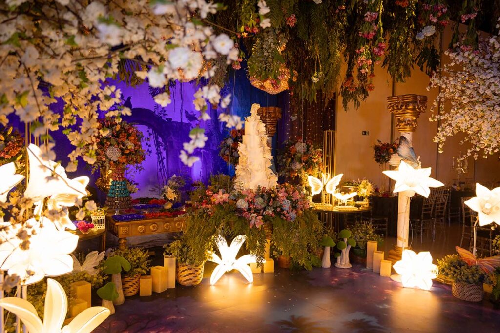 Cenário da festa com luminárias floridas, flores suspensas e bolo ao centro