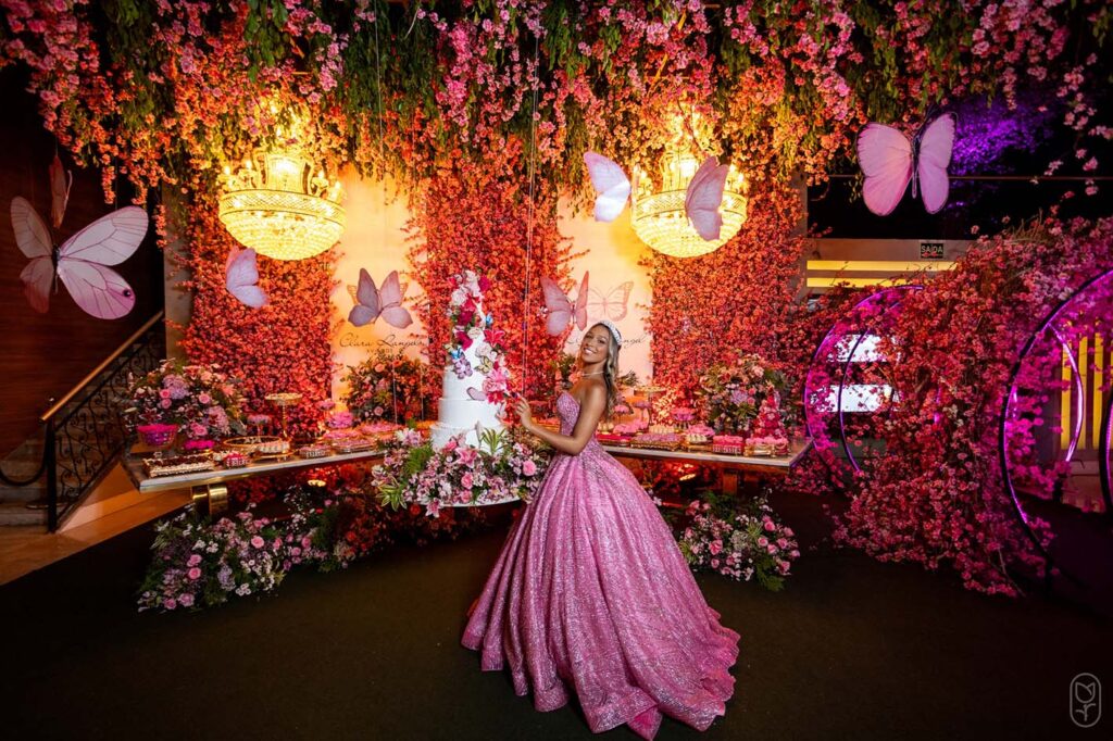 Debutante em vestido rosa diante do arco de flores e da mesa do bolo