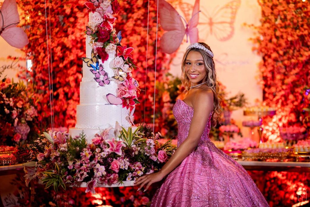 Debutante em vestido rosa ao lado do bolo cercado por flores