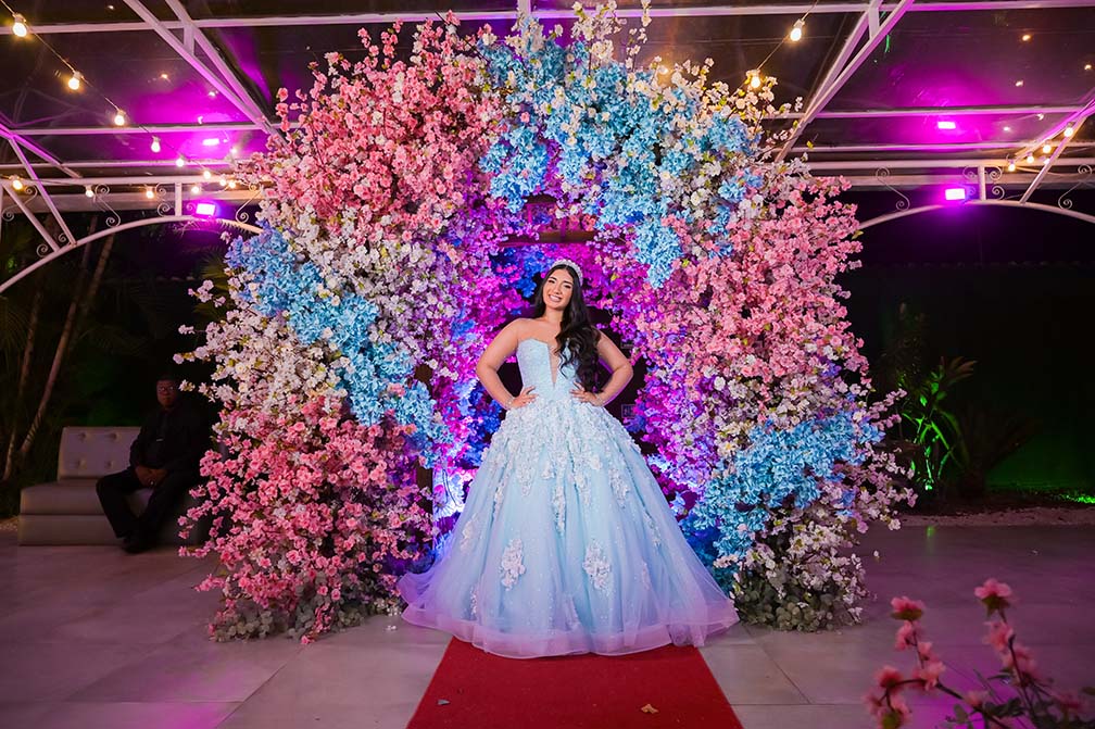Debutante em vestido azul sob o arco de flores em tons de rosa e azul