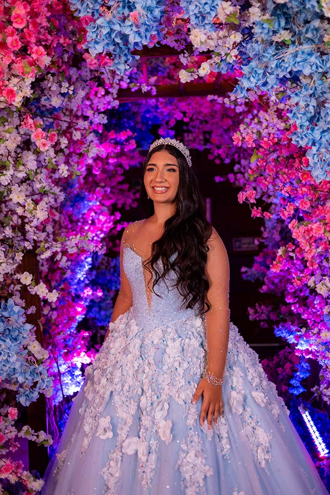 Debutante em vestido azul diante da parede de flores coloridas