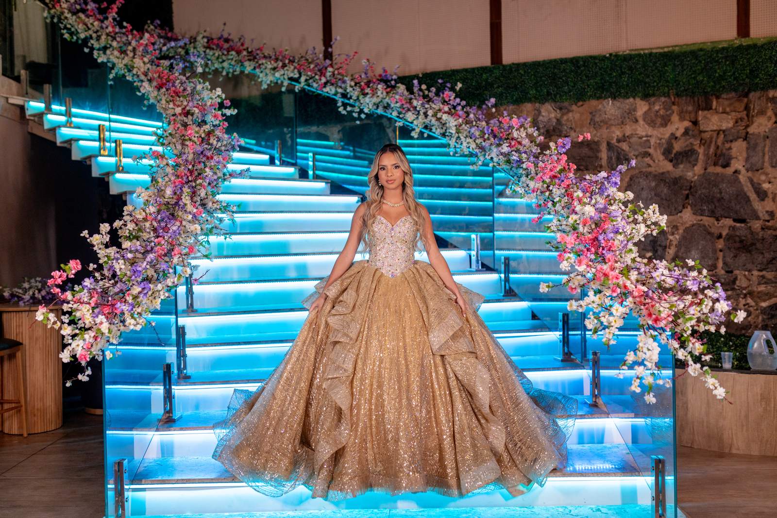 Debutante em vestido dourado na escadaria iluminada em azul com arco floral na festa de 15 anos tema Aladdin